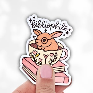 Peut inclure: Un autocollant représentant un lapin de dessin animé portant des lunettes, assis dans une tasse à thé sur deux livres roses. La tasse à thé est ornée de motifs floraux et le mot "bibliophile" est écrit au-dessus du lapin en cursive.