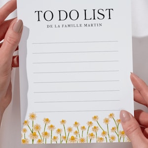 Peut inclure: Bloc-notes blanc "TO DO LIST DE LA FAMILLE MARTIN" avec texte noir. Le bloc-notes a des lignes horizontales pour l'écriture et une bordure florale de marguerites en bas.