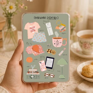 Peut inclure: Une planche d'autocollants rectangulaire vert sauge intitulée BOOKWORM ESSENTIALS présentant des illustrations d'un t-shirt rose, d'un short à motifs, d'une carte de bibliothèque, d'un porte-clés, d'un mug rose, d'un plaid, de biscuits, d'une citrouille, d'une coque de téléphone, d'une liseuse, d'une lampe et de lunettes.