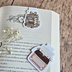 Peut inclure: Deux marque-pages magnétiques posés sur un livre ouvert. L'un représente une pile de cinq livres attachés par un ruban marron, une loupe et une clé. L'autre montre une machine à écrire vintage avec le texte once upon a time.