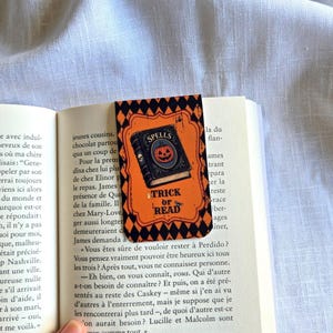 Peut inclure: Un marque-page rectangulaire avec un fond orange et une bordure à motif de losanges noirs. Le centre présente une illustration d'un livre bleu foncé intitulé SPELLS avec un motif de citrouille et le texte TRICK OR READ en lettres noires.