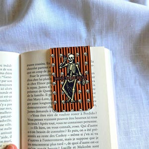 May include: A rectangular bookmark featuring a vertical orange and black striped pattern with a central illustration of a skeleton holding a book. The text BONE TO BE READ is printed in the centre, surrounded by small white skull icons throughout.