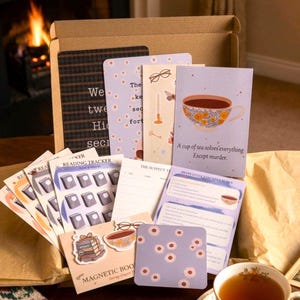Peut inclure: Une boîte en carton remplie d'articles de papeterie, notamment des suivis de lecture, des marque-pages et des cartes de vœux. Une carte présente une illustration de tasse à thé fleurie avec le texte, A cup of tea solves everything. Except murder.