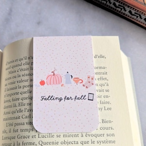 Peut inclure: Un marque-page rectangulaire avec un fond rose clair à petits points rouges. Le motif comprend une pomme rouge, une citrouille rose, une cafetière bleue, une tasse et des chaussettes. Le texte Falling for fall est imprimé en noir avec une icône de livre.