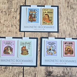 Peut inclure: Trois ensembles de deux marque-pages magnétiques sur des cartes blanches à bordures bleu foncé. Chaque marque-page présente une illustration en forme de timbre d'animaux lisant des livres, avec des textes comme Rainy Day Chapter Club, Library Card, Bedtime Chapter Club, Cozy Blanket Reading Club, Late Night Reader Club et Anti-Social Book Club.