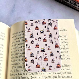 Peut inclure: Un marque-page rectangulaire sur fond beige clair orné d'un motif répétitif de petites illustrations sur le thème d'Halloween, incluant des citrouilles orange, des fantômes blancs, des chaudrons noirs et des silhouettes de maisons hantées.