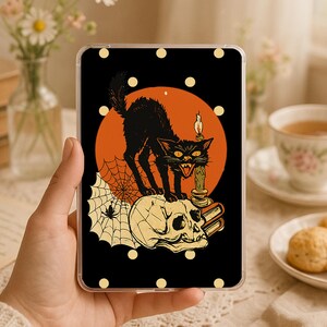 May include: A rectangular clear phone case featuring a black background with a graphic illustration of a black cat standing on a human skull next to a lit candle and a spider web, all set against a large orange circle with yellow polka dots.