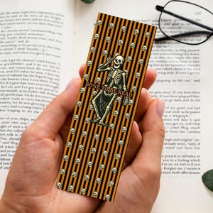 May include: A rectangular bookmark featuring a pattern of black and orange vertical stripes with small white skulls scattered throughout. In the centre, a skeleton illustration is positioned above the text BONE TO BE READ in black letters.