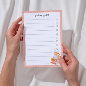 Peut inclure: Un bloc-notes rectangulaire avec une bordure pêche claire ornée d'un motif de citrouilles. Le centre blanc porte le texte "fall into reading" en haut, suivi de dix lignes horizontales avec des cases à cocher et une pile de livres avec un mug renard.