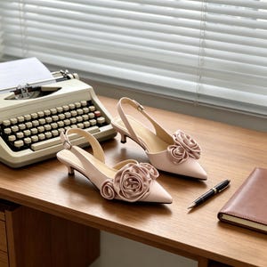 Puede incluir: Un par de zapatos de tacón rosa pálido con punta y tacón pequeño. Cada zapato tiene una gran rosa de tela en la punta. Los zapatos están sobre una superficie de madera, junto a una máquina de escribir vintage. Los tacones miden unos 5 cm.