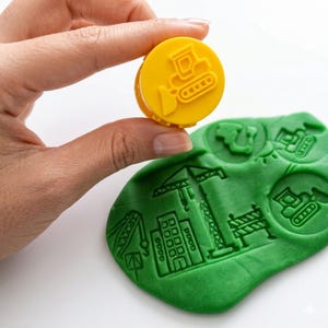 May include: A yellow construction-themed stamp held above green playdough. The stamp features a bulldozer design. The playdough has impressions of construction vehicles, buildings, and a crane. This set is designed for creative play.