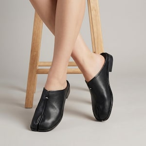 Puede incluir: Zapatos negros de cuero sin cordones con diseño de puntera dividida. Los zapatos tienen un tacón bajo y una estética minimalista. El calzado se muestra con una persona sentada en un taburete de madera.