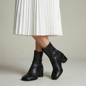 May include: Black leather ankle boots with a split toe design and a block heel. The boots have a sleek, minimalist style and are paired with a white pleated skirt. The boots are a versatile footwear choice.