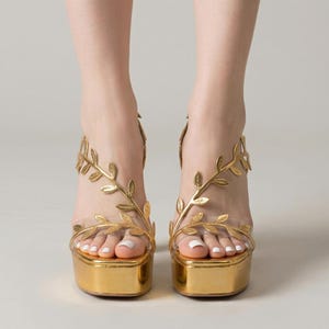 May include: Gold platform sandals with clear straps and leaf-shaped accents. The sandals have a square toe and a thick, metallic gold platform. The leaf design wraps around the ankle and foot, adding a decorative element to the footwear.