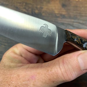 May include: A stainless steel knife with a sharp, metallic blade featuring an engraved bird icon near the bolster. The handle is constructed from a dark, textured material with a mottled brown and black pattern and visible metallic rivets.