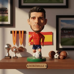 May include: A bobblehead figurine of a soccer player in a red jersey with the number 7, blue shorts, and a soccer ball at his feet. The figurine is on a green base with the text "BOBZBALLZ". The background includes a Spanish flag and framed pictures.