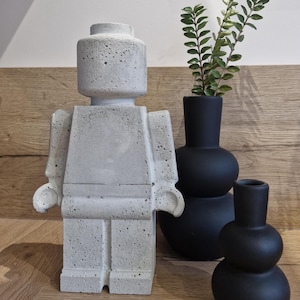 Puede incluir: Una gran figura de hormigón gris con forma de figura de juguete clásica se alza sobre una superficie de madera. Junto a ella, tres jarrones negros con acabado mate, uno de ellos con tallos verdes y frondosos. La figura tiene una superficie texturizada.