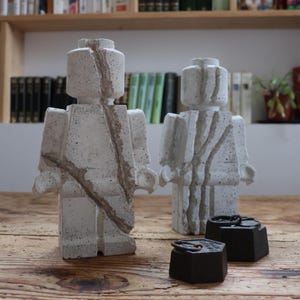 Puede incluir: Dos esculturas de hormigón con forma de figura de juguete clásica, con una combinación de colores gris claro y marrón. Dos pequeños objetos cuadrados de color marrón oscuro están delante de las esculturas. Las esculturas están sobre una superficie de madera.