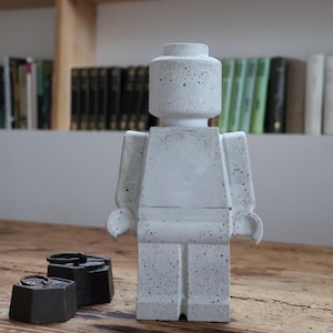 Può includere: Una statuetta in cemento grigio chiaro a forma di personaggio giocattolo squadrato con un perno cilindrico sulla testa. La superficie presenta una texture maculata. Accanto alla statuetta ci sono due pesi in metallo scuro con piccoli anelli.