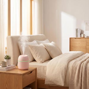 Pode incluir: Um difusor de óleos essenciais rosa com uma luz verde está em uma mesa de cabeceira de madeira ao lado de uma cama com roupa de cama creme. O difusor está emitindo vapor. Uma pequena planta em vaso e uma caneca também estão na mesa.