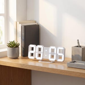 May include: A white digital clock showing 20:05, with illuminated white LED numerals. The clock is on a wooden shelf, alongside a small succulent in a grey pot, books, and a pen holder. The clock has a contemporary, minimalist design.