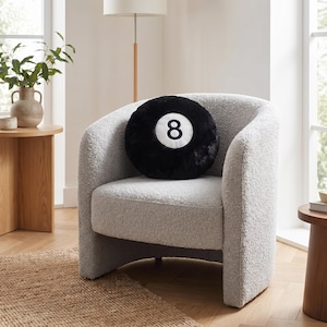 May include: A black, circular cushion featuring the number 8 on a white background, placed on a light grey, curved armchair. The cushion is made of a soft, plush material. The chair sits on a jute rug.
