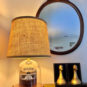 Puede incluir: Una lámpara de mesa de estilo vintage con una pantalla de tela beige y una base de madera. La lámpara presenta una cúpula de cristal transparente que muestra un instrumento mecánico. Un espejo ovalado de madera oscura cuelga en la pared. Dos sujetalibros dorados con forma de pato están sobre una mesa.