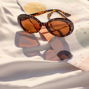 May include: Oval-shaped sunglasses with a tortoiseshell pattern. The sunglasses have brown-tinted lenses and are resting on a white fabric with a colorful design. The image is well-lit, highlighting the sunglasses' details.