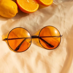 Puede incluir: Gafas de sol redondas naranjas con montura negra sobre una tela color crema, junto a naranjas en rodajas y un limón entero. Las gafas de sol tienen un estilo retro, y las lentes son de color naranja vibrante.