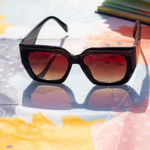 May include: Black square-framed sunglasses with gradient brown lenses. The sunglasses are set against a colorful abstract background. The frames are thick and have a glossy finish, and the lenses transition from a darker brown at the top to a lighter shade at the bottom.