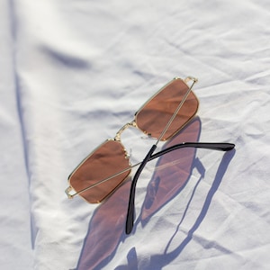 May include: Gold-framed sunglasses with rectangular lenses in a soft, peach hue. The black temples are extended, casting a shadow on the white fabric. The sunglasses are a stylish accessory.