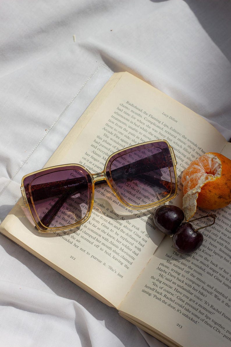 Puede incluir: Gafas de sol cuadradas con lentes moradas descansan sobre un libro abierto. Las gafas tienen una montura dorada. Dos cerezas y una naranja pelada est&aacute;n al lado del libro. La escena est&aacute; iluminada por luz natural.