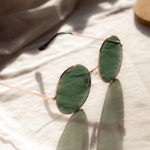 May include: Round sunglasses with gold-colored frames and green lenses. The sunglasses are resting on a white surface, with their shadow visible. The image is well-lit, highlighting the details of the eyewear.