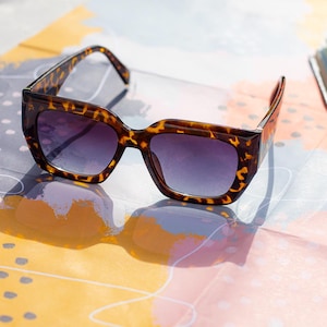 May include: Square-framed sunglasses with a tortoiseshell pattern. The lenses are a gradient of purple to dark blue. The sunglasses are resting on a colorful abstract patterned surface with yellow, orange, and blue hues.