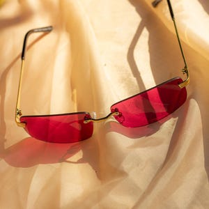 Puede incluir: Gafas de sol sin montura con montura dorada y lentes rojas. Las gafas de sol tienen forma rectangular y están sobre una tela beige.