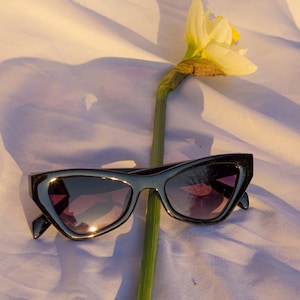 Könnte beinhalten: Schwarze Cat-Eye-Sonnenbrille mit violett getönten Gläsern liegt auf einer weißen Oberfläche neben einer Narzisse. Die Sonnenbrille hat ein schlankes, modernes Design. Die Narzisse hat eine gelbe Mitte und weiße Blütenblätter.