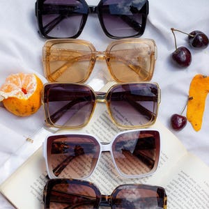 Puede incluir: Una variedad de gafas de sol cuadradas en negro, &aacute;mbar, marr&oacute;n, blanco y carey. Las gafas de sol est&aacute;n dispuestas sobre un libro abierto con una naranja cortada y cerezas.