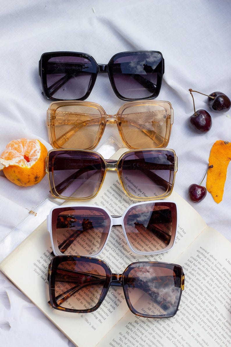 Puede incluir: Una variedad de gafas de sol cuadradas en negro, &aacute;mbar, marr&oacute;n, blanco y carey. Las gafas de sol est&aacute;n dispuestas sobre un libro abierto con una naranja cortada y cerezas.