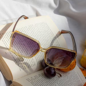 Puede incluir: Gafas de sol cuadradas con montura marr&oacute;n claro y lentes moradas sobre un libro abierto. Las gafas est&aacute;n junto a unas cerezas y una peque&ntilde;a naranja. Las p&aacute;ginas del libro est&aacute;n amarillentas.