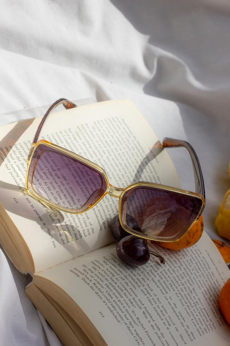 Puede incluir: Gafas de sol cuadradas con montura marr&oacute;n claro y lentes moradas sobre un libro abierto. Las gafas est&aacute;n junto a unas cerezas y una peque&ntilde;a naranja. Las p&aacute;ginas del libro est&aacute;n amarillentas.