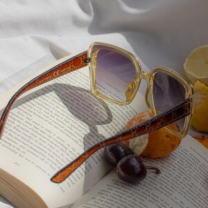 Puede incluir: Gafas de sol cuadradas con lentes degradadas moradas sobre un libro abierto, con fruta. Las monturas son de color amarillo claro con un dise&ntilde;o marr&oacute;n en las patillas. Las p&aacute;ginas del libro son visibles.