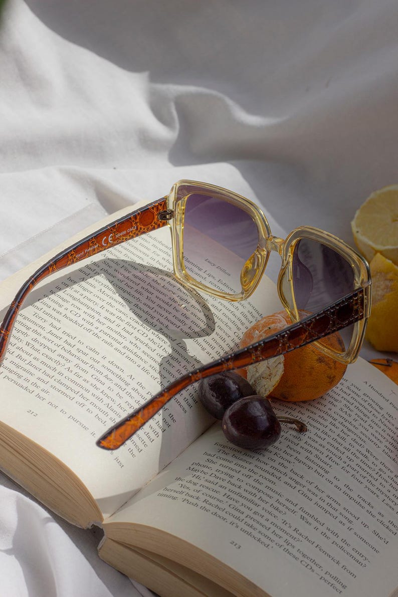 Puede incluir: Gafas de sol cuadradas con lentes degradadas moradas sobre un libro abierto, con fruta. Las monturas son de color amarillo claro con un dise&ntilde;o marr&oacute;n en las patillas. Las p&aacute;ginas del libro son visibles.