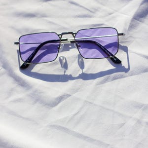 Puede incluir: Gafas de sol rectangulares con montura plateada y lentes moradas. El diseño es moderno, con una fina montura plateada y lentes moradas oscuras. La imagen está bien iluminada, destacando el diseño elegante de las gafas.