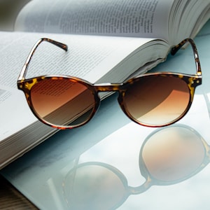 May include: Round sunglasses with a tortoiseshell pattern frame and gradient brown lenses sit on an open book. The sunglasses are reflected on a light blue surface. The book's pages are visible, suggesting a reading theme.