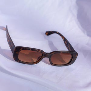 Può includere: Occhiali da sole rettangolari con montatura a fantasia tartarugata e lenti marroni. Gli occhiali da sole sono appoggiati su uno sfondo bianco. Anche le aste degli occhiali da sole hanno un motivo tartarugato.