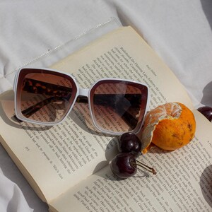 May include: White-framed sunglasses with brown lenses rest on an open book. A peeled orange and cherries sit nearby. The book's pages display text, creating a relaxed, summery scene.