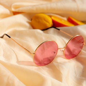 Puede incluir: Gafas de sol rosas redondas con montura dorada y patillas negras sobre tela crema. Un limón y rodajas de naranja en el fondo. Diseño retro y elegante.