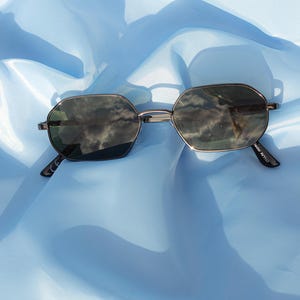 May include: Silver-framed sunglasses with dark lenses. The glasses have a geometric, octagonal shape. The temples are thin and silver. The sunglasses are set against a light blue, silky background.