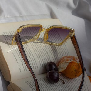 Puede incluir: Gafas de sol cuadradas transparentes con degradado p&uacute;rpura, apoyadas en un libro abierto. Las monturas son amarillas transl&uacute;cidas con patillas marrones estampadas. Cerezas y una mandarina junto al libro.