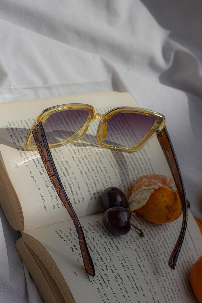 Puede incluir: Gafas de sol cuadradas transparentes con degradado p&uacute;rpura, apoyadas en un libro abierto. Las monturas son amarillas transl&uacute;cidas con patillas marrones estampadas. Cerezas y una mandarina junto al libro.